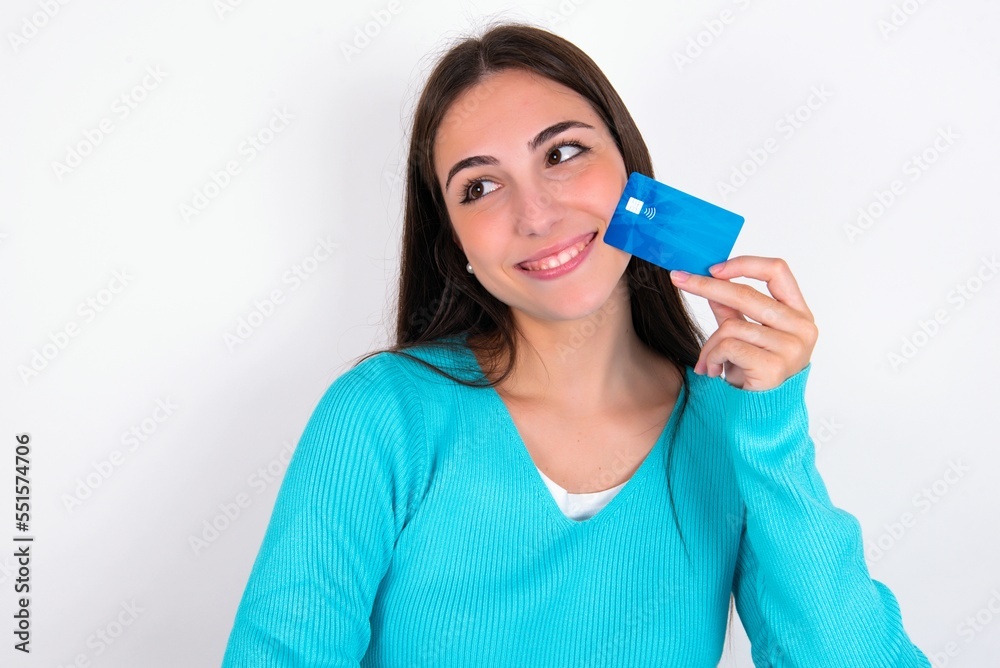 Lovely Young caucasian woman wearing blue sweater over white background showing credit card and looking away at copy space. Pensive Lovely Young caucasian woman wearing blue sweater over white background showing credit card and looking away at copy space. Pensive
