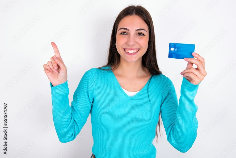Smiling Young caucasian woman wearing blue sweater over white background showing debit card pointing finger empty space Smiling Young caucasian woman wearing blue sweater over white background showing debit card pointing finger empty space