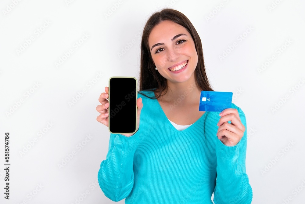 Young caucasian woman wearing blue sweater over white background opened bank account, holding smartphone and credit card, smiling, recommend use online shopping application Young caucasian woman wearing blue sweater over white background opened bank account, holding smartphone and credit card, smiling, recommend use online shopping application