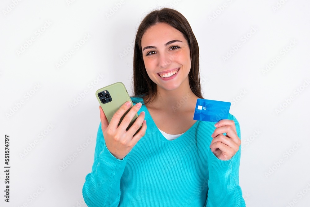 Young caucasian woman wearing blue sweater over white background wears stereo headphones on ears holds modern mobile phone and credit card Young caucasian woman wearing blue sweater over white background wears stereo headphones on ears holds modern mobile phone and credit card