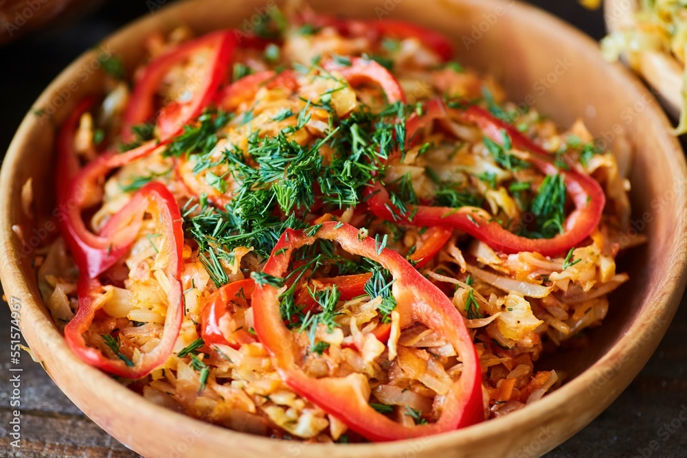 Fried cabbage in a wooden bowl on the table. Decoration of sweet pepper and green dill. Fried cabbage in a wooden bowl on the table. Decoration of sweet pepper and green dill.
