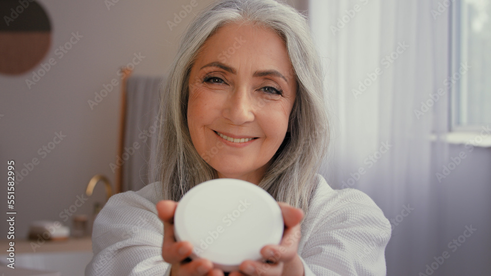 Happy old Caucasian 50s senior model gray-haired mature woman smile toothy 60s lady in bathroom showing jar of anti-aging cosmetic natural organic sunscreen mineral cream moisturizing face skin care Happy old Caucasian 50s senior model gray-haired mature woman smile toothy 60s lady in bathroom showing jar of anti-aging cosmetic natural organic sunscreen mineral cream moisturizing face skin care