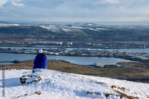 Woman listening to music and loking at the horizon on the snow. Person sitting and chilling on the mountain. Views over Belfast harbor from Cave Hill in winter. Northern Ireland.