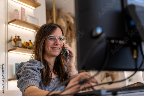 Positive adult female employee with long dark hair in casual wear and eyeglasses smiling and typing on computer keyboard while having phone call 