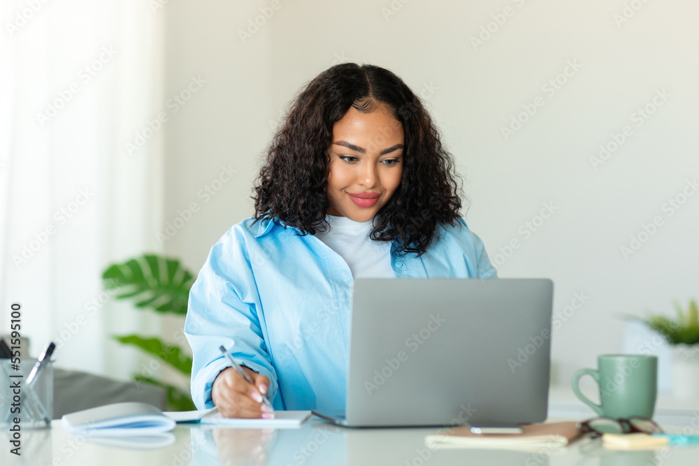 Distance education. Black obese woman sitting at desk, using laptop and writing in notebook, studying online at home Distance education. Black obese woman sitting at desk, using laptop and writing in notebook, studying online at home