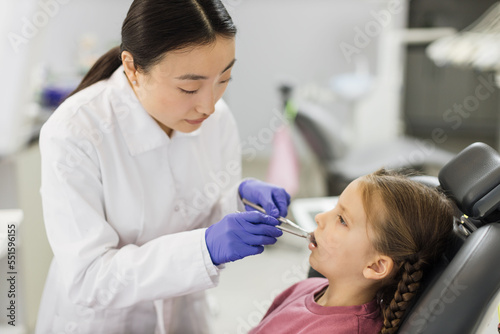 Side view of smiling pretty asian doctor in white uniform and gloves  making examination or curing caries and toothache for her little patient  cute kid girl  sitting in dental chair.