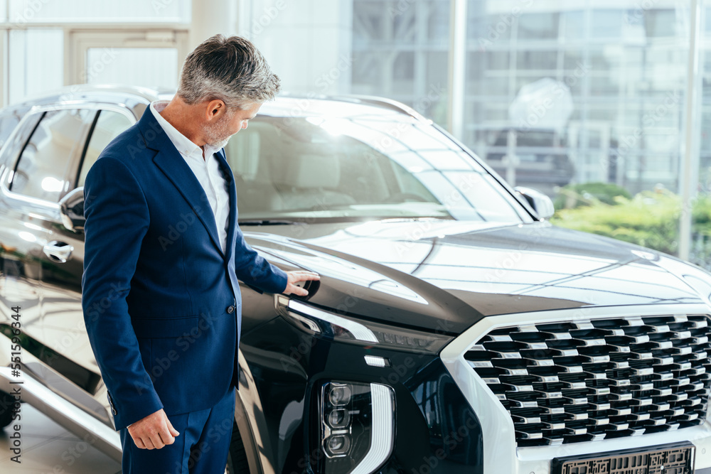 Making his choice. Serious mature grey hair beard businessman salesman wearing blue formal suit standing next to new car in showroom using his smartphone.