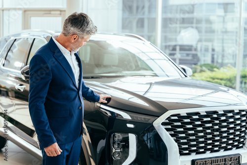 Making his choice. Serious mature grey hair beard businessman salesman wearing blue formal suit standing next to new car in showroom using his smartphone. © Iryna