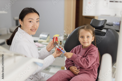 Happy smiling asian female dentist tells little child girl how to brush the teeth on artificial jaw model. Caries prevention  pediatric dentistry  milk teeth hygiene concept.