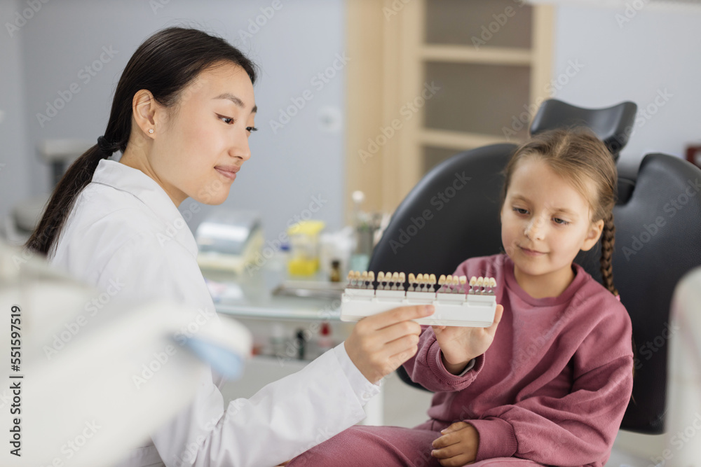 Happy young smiling preschool girl in casual clothes in the dentist light modern office. Asian female doctor in white coat makes comparison of the patient teeth with the dental whitening chart. Happy young smiling preschool girl in casual clothes in the dentist light modern office. Asian female doctor in white coat makes comparison of the patient teeth with the dental whitening chart.