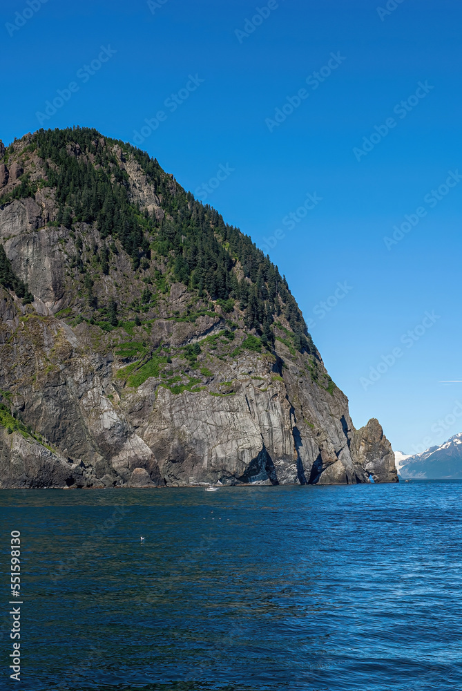 Resurrection Bay, Alaska, USA - July 22, 2011: Steep gray rocky cliff descends into blue water and appear to have formed a drinking lion's head its mouth submerged. Mountain with snow patches