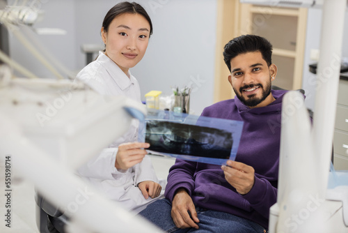 Attractive young bearded man  sitting on dentist chair and looking at x-ray picture of scan image of teeth together with his cheerful female asian dentist at clinic.