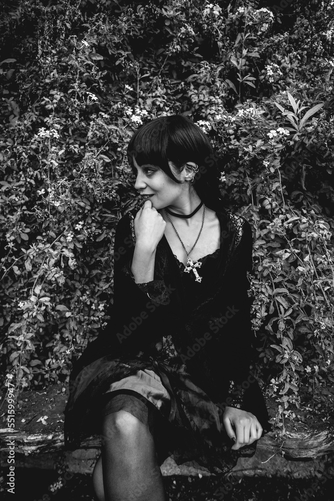 Young and skinny hispanic goth girl with black dress and cross seated in a rustic wood bench next to white flowers in the forest (in black and white) Young and skinny hispanic goth girl with black dress and cross seated in a rustic wood bench next to white flowers in the forest (in black and white)