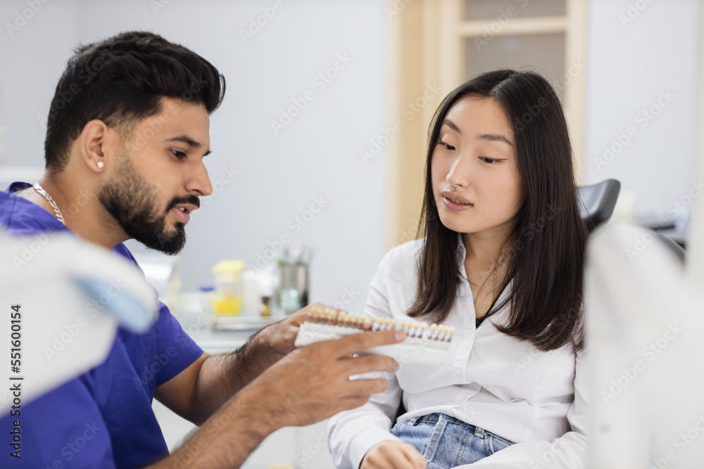 Attractive male dentist checking and selecting color of young asian woman teeth. Visual method of subjective perception color of filling by a doctor. Dentist choosing color of filling from palette Attractive male dentist checking and selecting color of young asian woman teeth. Visual method of subjective perception color of filling by a doctor. Dentist choosing color of filling from palette