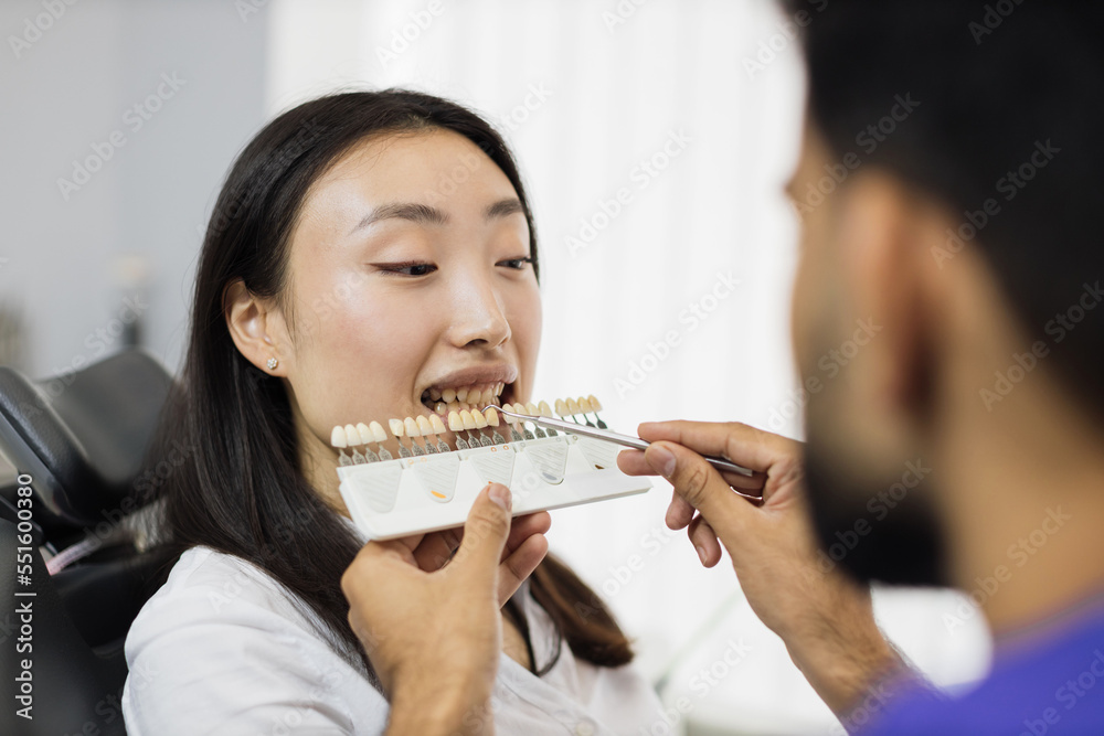 Dentist choosing color of filling from palette. Back view of male dentist checking and selecting color of young asian woman teeth indoors in modern hospital. Dentist choosing color of filling from palette. Back view of male dentist checking and selecting color of young asian woman teeth indoors in modern hospital.