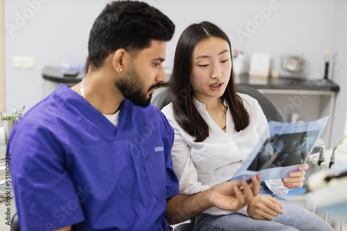 Attractive young asian woman  sitting on dentist chair and looking at x-ray picture of scan image of teeth together with her confident professional male dentist at clinic.