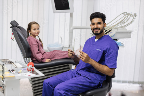 Children's teeth treatment. Smiling little girl sitting in the dentist's chair in light modern dental clinic to excited confident man dentist and looking at camera.