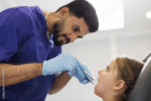 Side low angle view of smiling male professional doctor in blue uniform and gloves, making examination or curing caries and toothache for his little patient, cute kid girl, sitting in dental chair.
