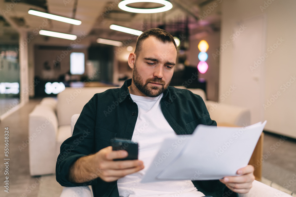 Fototapeta premium Serious businessman working with papers while using smartphone