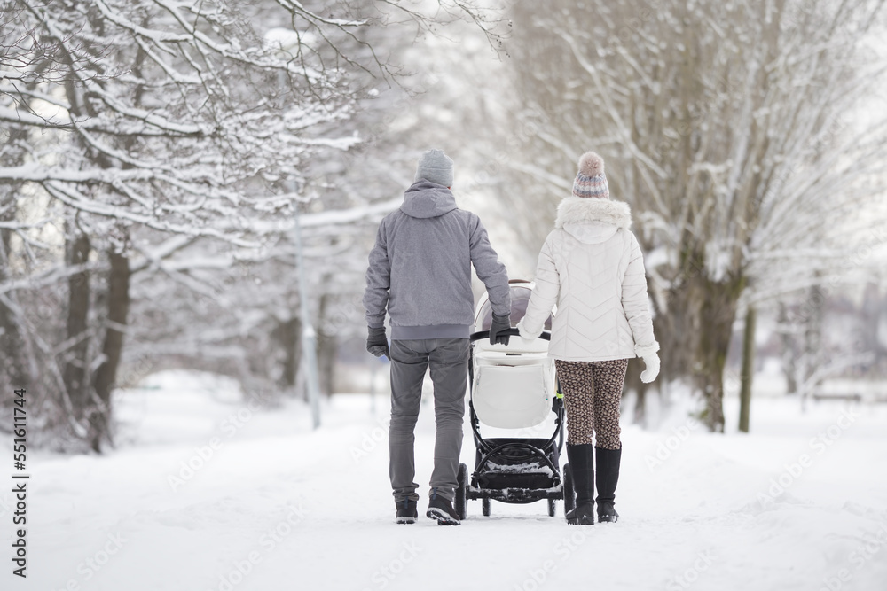 Young family pushing white baby stroller and walking on snow covered sidewalk at park in cold winter day. Enjoying peaceful stroll. Spending time with newborn and breathing fresh air. Back view. - obrazy, fototapety, plakaty 