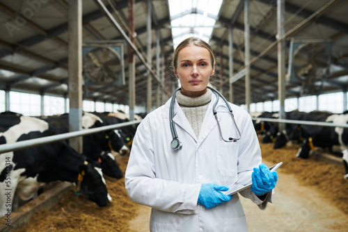 Young confident female veterinarian in whitecoat and gloves holding digital tablet and looking at camera against long cowsheds