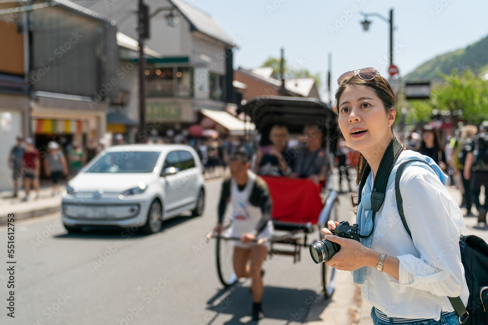 cheerful asian Japanese woman visitor looking at streetscape on sidewalk of Arashiyama Mountains street in Kyoto japan with people taking rickshaw ride at background on sunny day