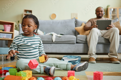 Full length portrait of cute black baby playing with toys on floor  father using computer in background  copy space