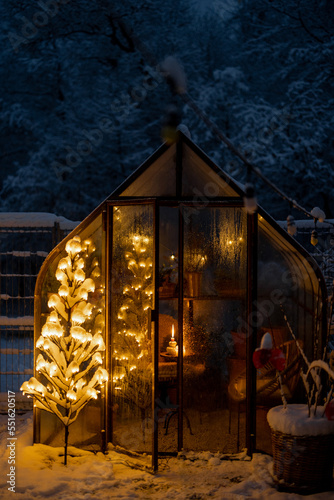 Beautiful snowy yard with vintage greenhouse, glowing tree garland and candle at dusk. Concept of New Year holidays and winter magic