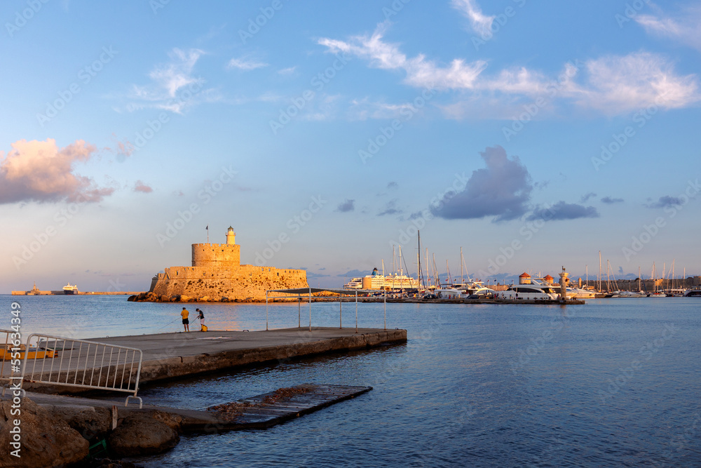 Obraz premium Colorful panoramic view of Agios Nikolaos fortress with yachts in Mandraki port at sunset, Rhodes Greece. High quality photo
