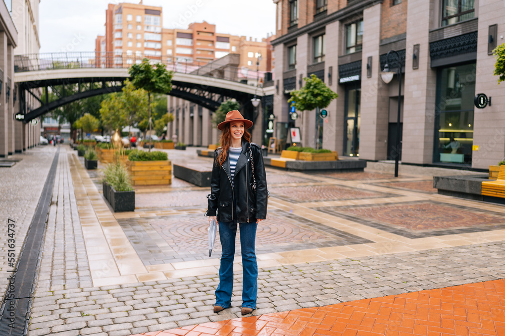 Full length portrait of happy redhead young woman wearing elegant hat standing with folded transparent umbrella on beautiful city street, smiling looking away. Concept of female lifestyle.