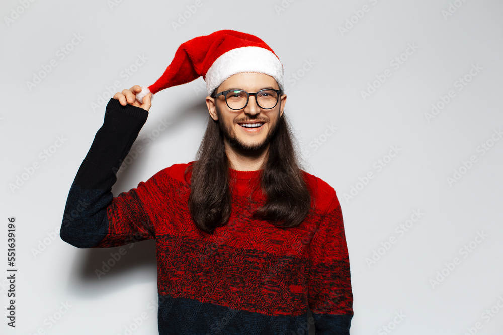 Studio portrait of young smiling man with long hair and eyeglasses, wears red Christmas sweater, holding pompon of Santa hat on a white background. Studio portrait of young smiling man with long hair and eyeglasses, wears red Christmas sweater, holding pompon of Santa hat on a white background.
