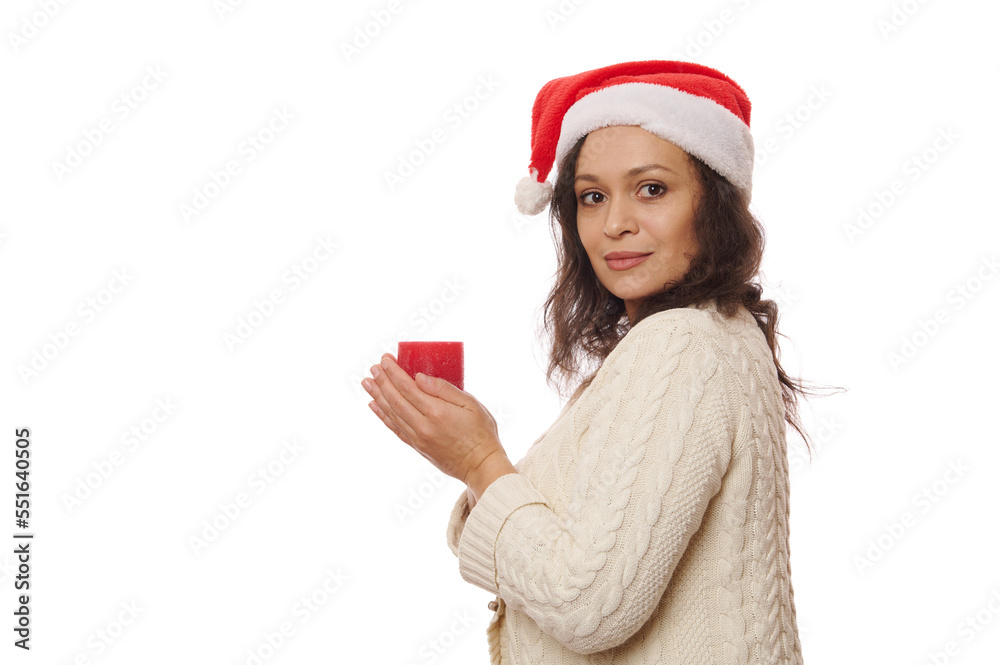 Side portrait of a Latin American pretty woman in Santa hat and beige sweater, holding a lit red Christmas candle and looking at camera through her shoulder, isolated over white background. Copy space Side portrait of a Latin American pretty woman in Santa hat and beige sweater, holding a lit red Christmas candle and looking at camera through her shoulder, isolated over white background. Copy space