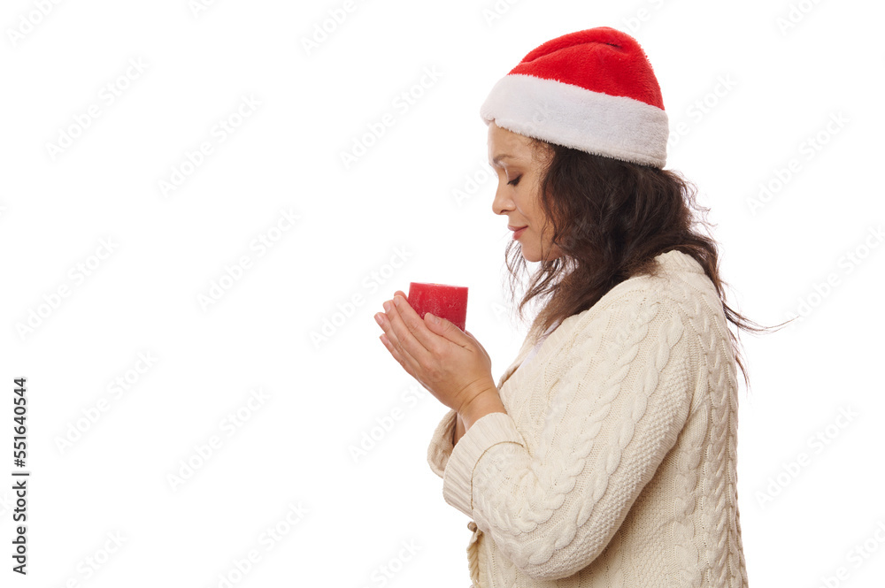 Side view of a beautiful serene woman in Santa hat and beige sweater, holding a red lit candle and makes wish for Christmas, isolated over white background. New Years concept. Copy advertising space Side view of a beautiful serene woman in Santa hat and beige sweater, holding a red lit candle and makes wish for Christmas, isolated over white background. New Years concept. Copy advertising space