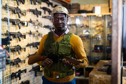 African-american man wearing armor vest and helmet while standing with machine gun in airsoft shop. © JackF