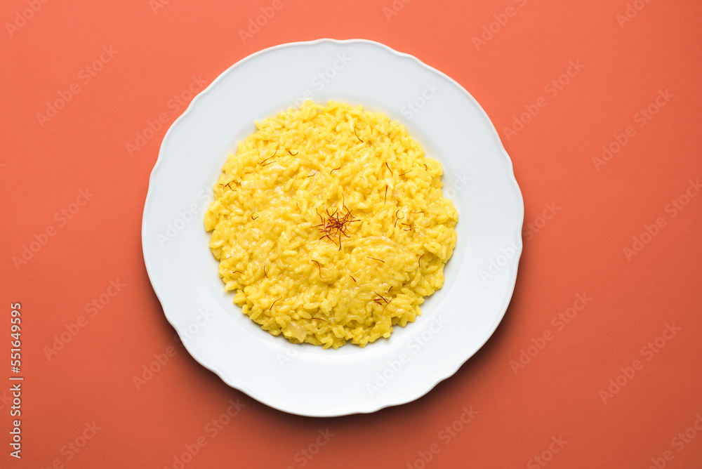 Risotto alla Milanese in a white aristocratic dish on the red terracotta background. A classic recipe for Lombard risotto with saffron for the cold season. Cuisine of the north Italy Risotto alla Milanese in a white aristocratic dish on the red terracotta background. A classic recipe for Lombard risotto with saffron for the cold season. Cuisine of the north Italy