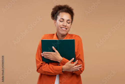Smiling African American woman with closed eyes holding book, love reading isolated on beige background 