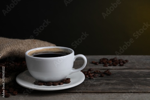 Cup of hot aromatic coffee and roasted beans on wooden table against dark background  space for text