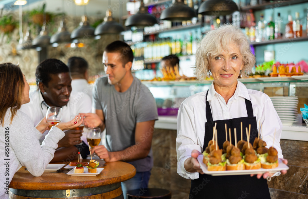 View on the European elder woman holding pinchos at a bar with diverse group in the background..