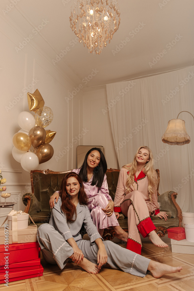 Three young caucasian interracial girlfriends celebrate birthday in room with gifts. Long-haired ladies wear nightgown and pants. Concept of spending time together