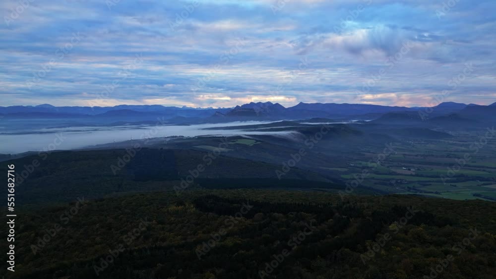 The morning mist trapped in the valley flows to the other side of the mountain. Panoramic view of the French Alps in the morning. Wind power station in thick fog in the distance The morning mist trapped in the valley flows to the other side of the mountain. Panoramic view of the French Alps in the morning. Wind power station in thick fog in the distance