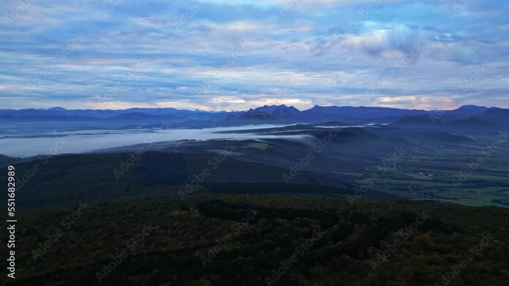 The morning mist trapped in the valley flows to the other side of the mountain. Panoramic view of the French Alps in the morning. Wind power station in thick fog in the distance The morning mist trapped in the valley flows to the other side of the mountain. Panoramic view of the French Alps in the morning. Wind power station in thick fog in the distance