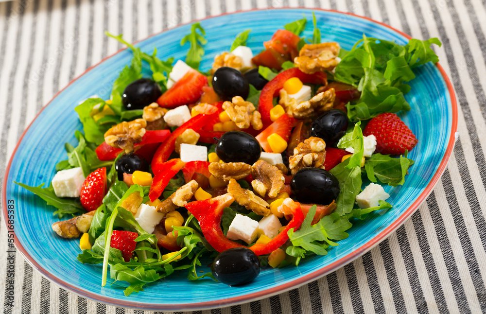 Fresh green arugula salad with vegetables, goat cheese and ripe strawberry decorated with black olives, walnuts and corn kernels