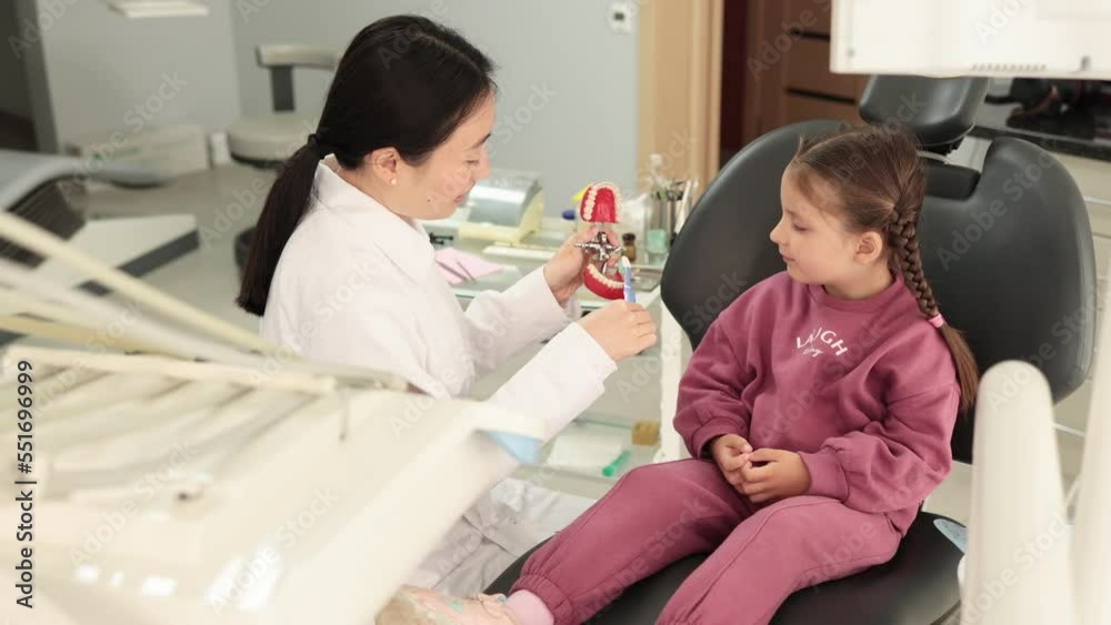 Smiling happy child girl with sitting at the dental office and looking at brush, while female young dentist shows how to brush teeth on artificial jaw model. Smiling happy child girl with sitting at the dental office and looking at brush, while female young dentist shows how to brush teeth on artificial jaw model.