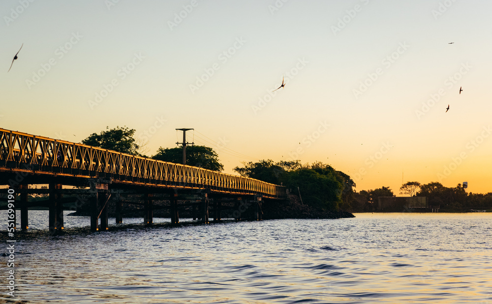 Beautiful landscape with an old iron and wooden bridge, a river and birds flying in the sky during sunset in Colonia Carlos Pellegrini, Iberá Wetland Provincial Nature Reserve, Corrientes, Argentina.