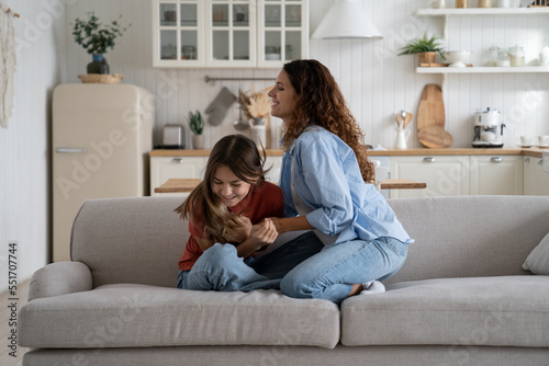 Overjoyed mother enjoying weekend with teen daughter, happy loving mom tickling kid, sitting together on sofa in living room, teenage girl playing with mommy. Fun family activities at home