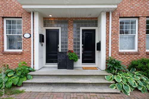 Two black metal doors with a black dropbox on the step of duplex houses. It has a red brick wall with white wooden trim. There are double hung windows on both sides of the entrance.