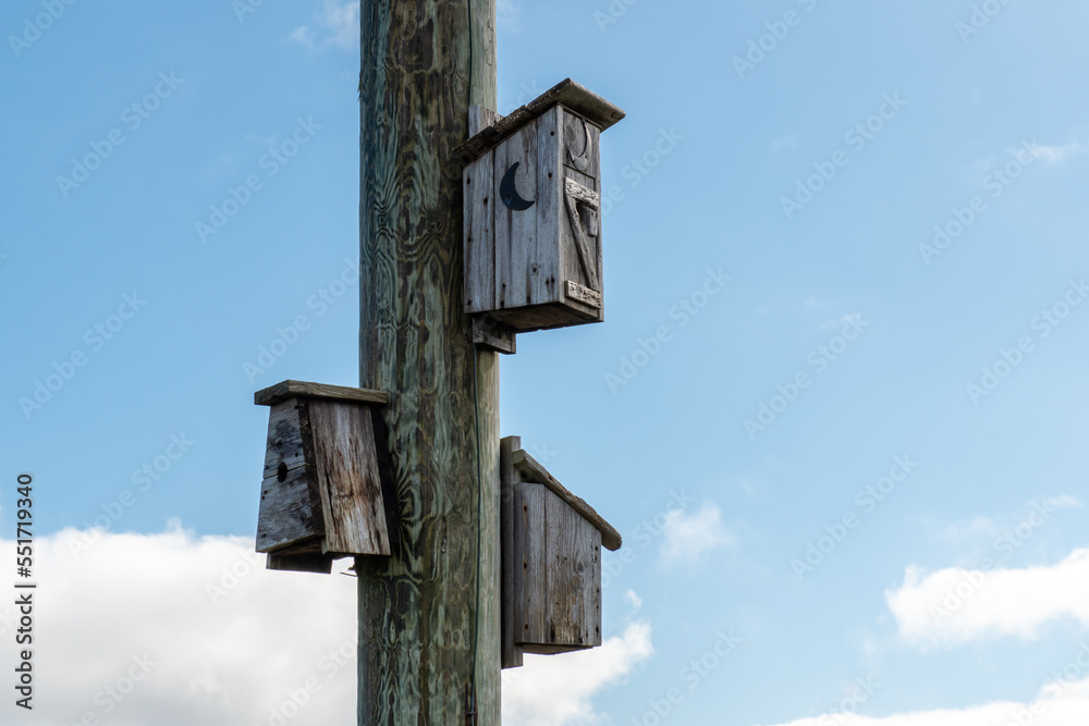 A post with three large wooden birdhouses for small birds. The birdhouses have single round entry holes in the center of the structures. One has a half moon shape opening. The wood is worn and grey. A post with three large wooden birdhouses for small birds. The birdhouses have single round entry holes in the center of the structures. One has a half moon shape opening. The wood is worn and grey.