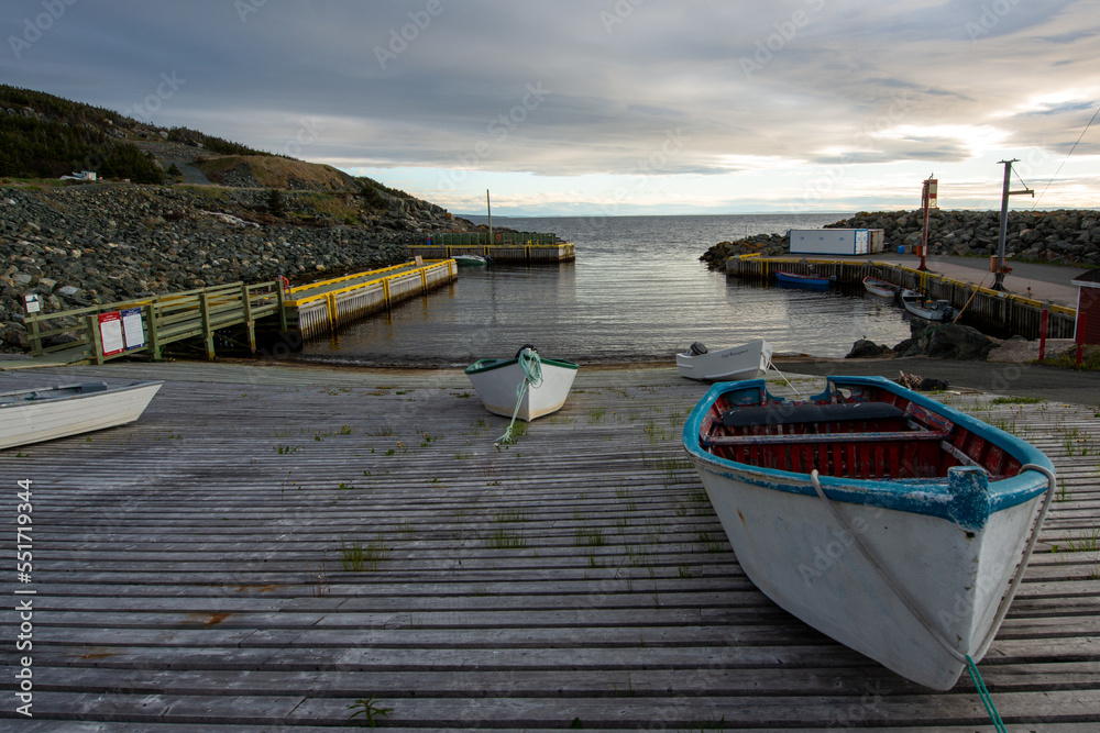 Naklejka premium Multiple small white wooden and fiberglass open fishing boats tied to a slipway in a small sheltered fishing harbor. The sky is cloudy and grey with yellow and orange hues from the setting sun.