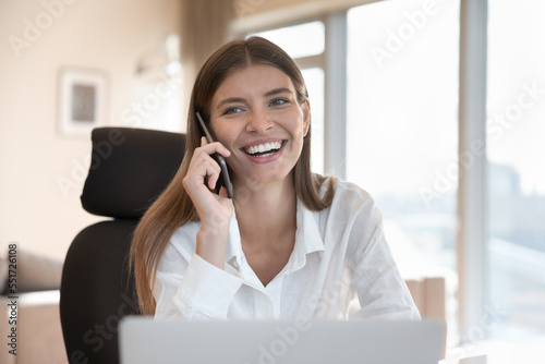 Cheerful pretty freelance employee girl using gadgets at workplace table, enjoying business communication, working from home, talking on mobile phone at laptop, smiling, laughing © fizkes