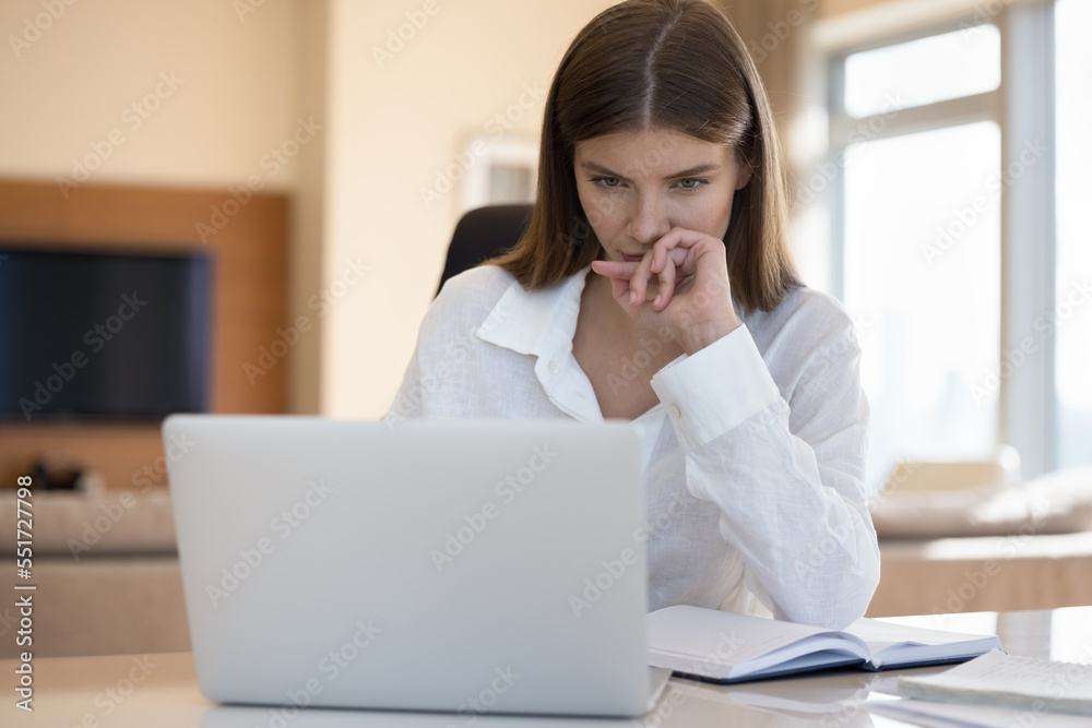 Serious thoughtful freelance employee woman sitting at home workplace table, looking at laptop screen, touching chin, face, reading text, watching online content, thinking on project Serious thoughtful freelance employee woman sitting at home workplace table, looking at laptop screen, touching chin, face, reading text, watching online content, thinking on project